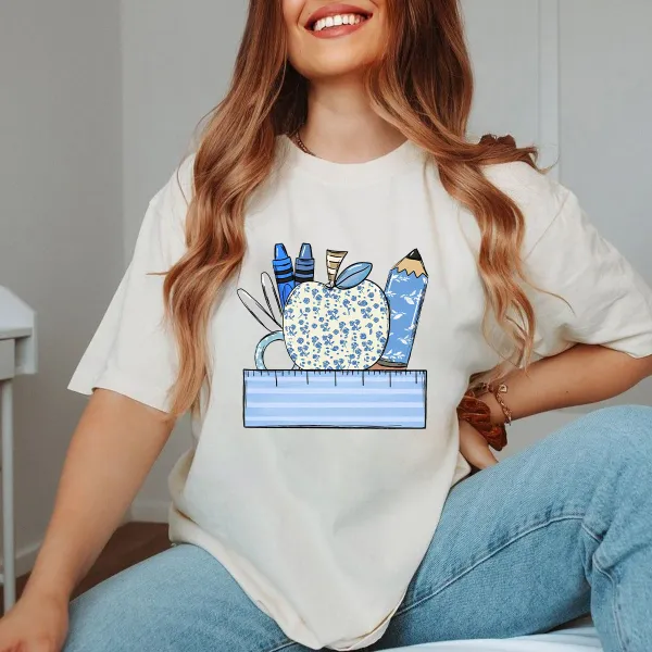 An Apple With A Blue Flower Pattern Surrounded By Stationery, Including Blue Crayons, Pencils, Scissors And Paper Clips, And A Light Blue Ruler At The Bottom, With A Casual T-Shirt.
